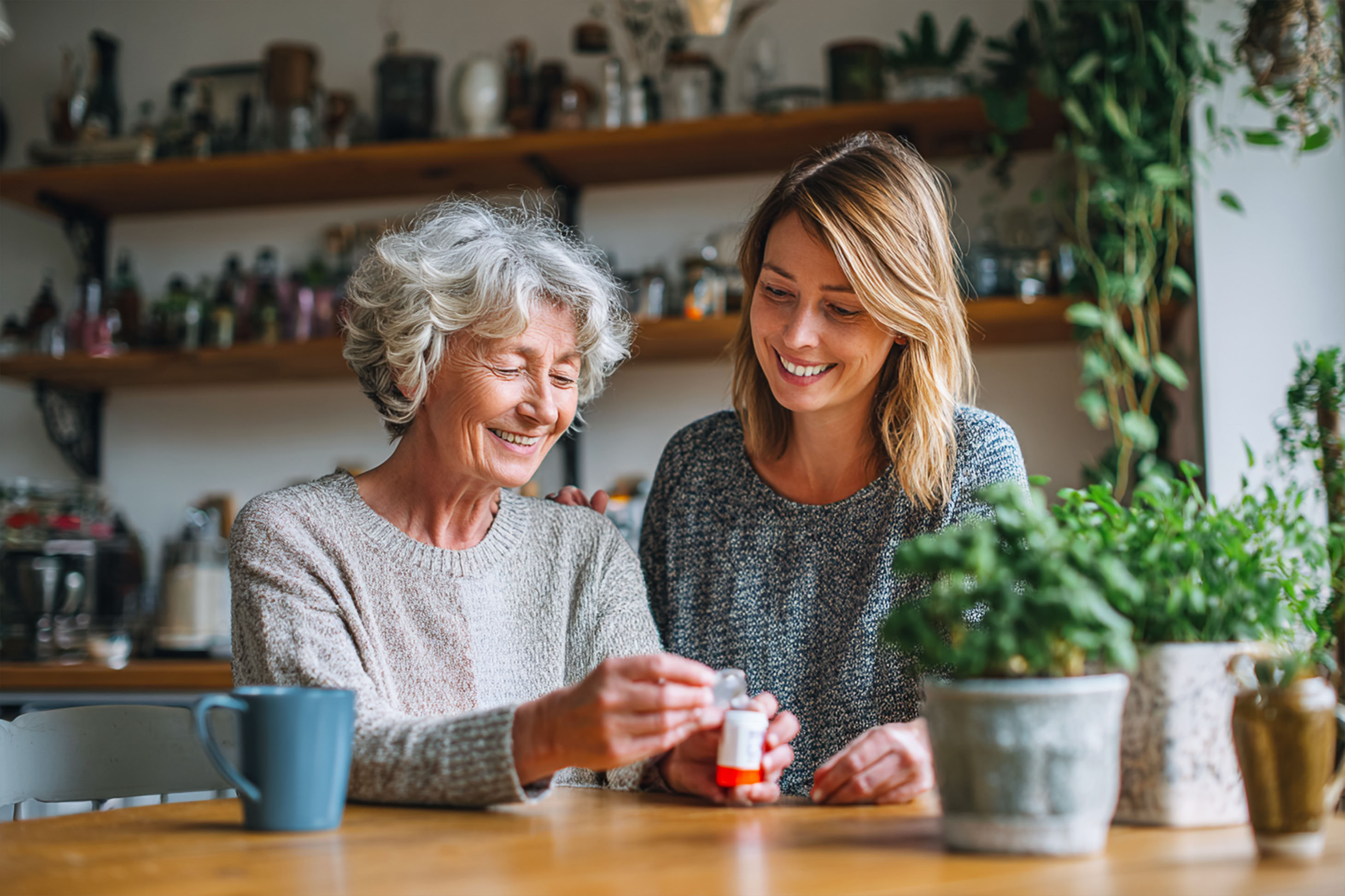 Elderly woman taking medicine at home with her daughter standing beside her in the kitchen.
