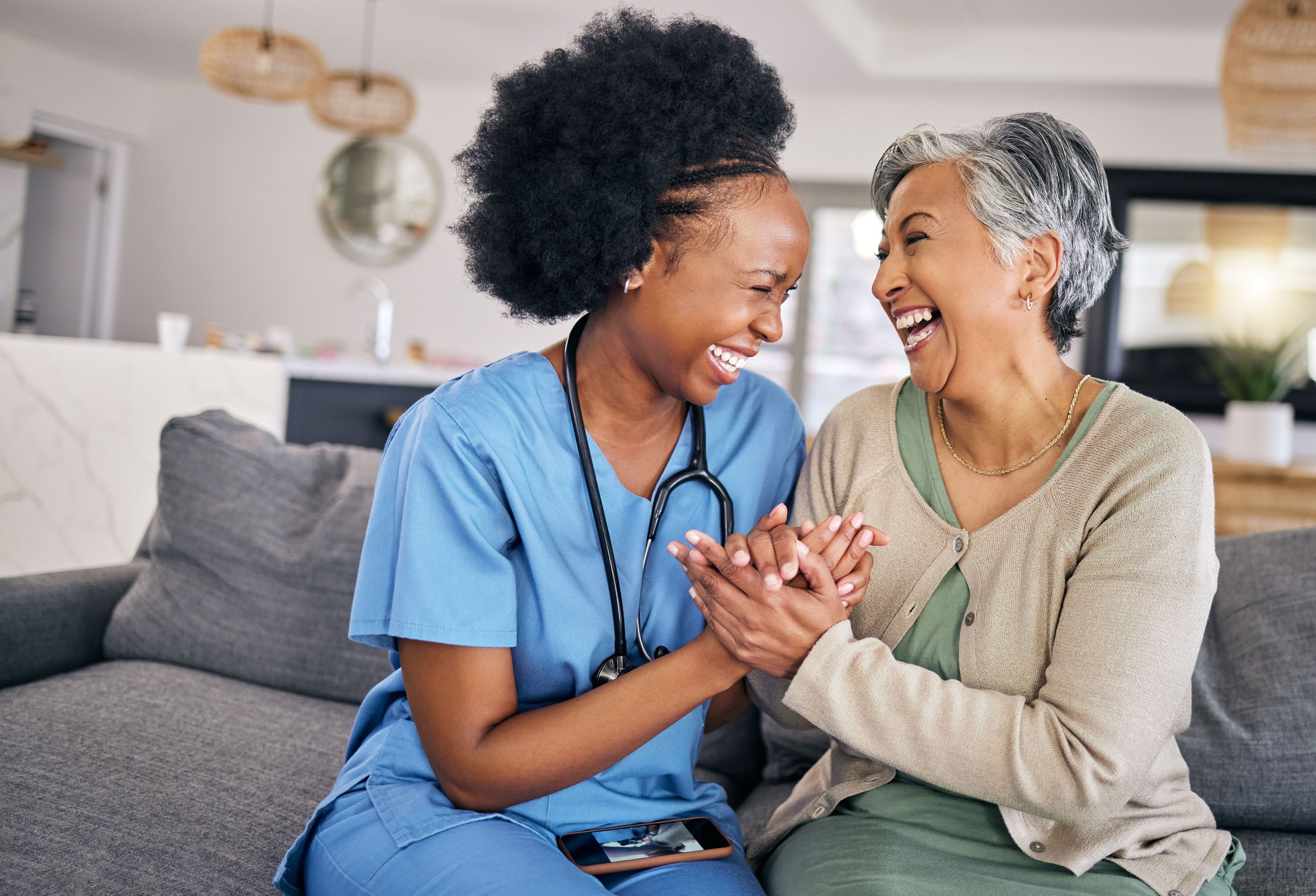 A nurse in blue scrubs and a stethoscope holds hands and shares a laugh with an older woman in a beige cardigan, sitting on a couch in a bright room.