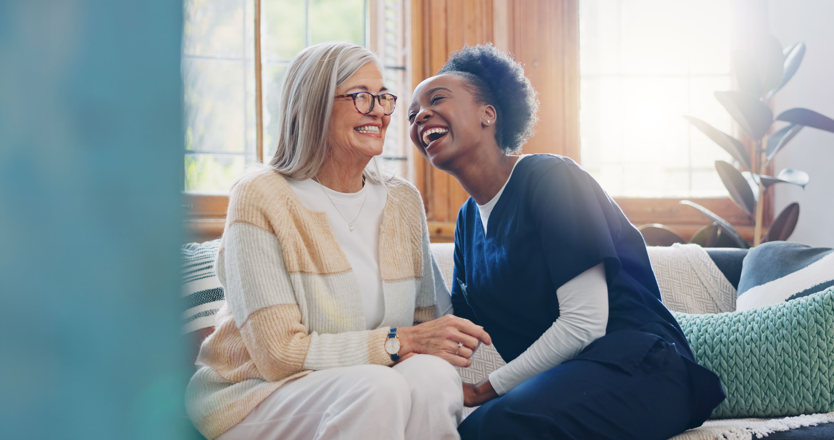 Two people sitting on a couch, one wearing a light-colored sweater and glasses, the other in dark scrubs, both smiling and holding hands.